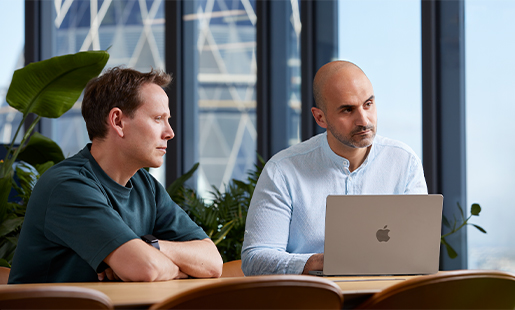 Ahmed, con su MacBook delante, sentado en una mesa con un colega y mirando a alguien fuera de cuadro.