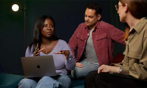 Ginelle con una MacBook en el regazo, sentada y trabajando con dos personas.