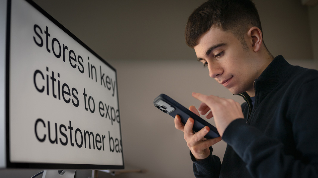 Un estudiante usa un iPhone mientras está sentado frente a una pantalla con el contenido de su clase de negocios en texto grande y ampliado