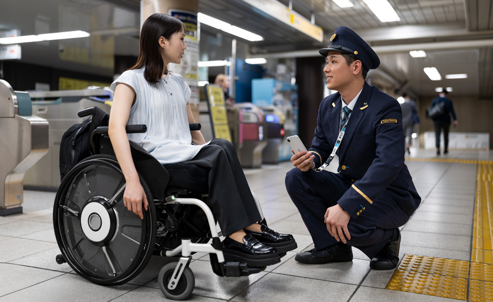 Una donna vestita in modo professionale su sedia a rotelle e un addetto della stazione che parlano sulla piattaforma.