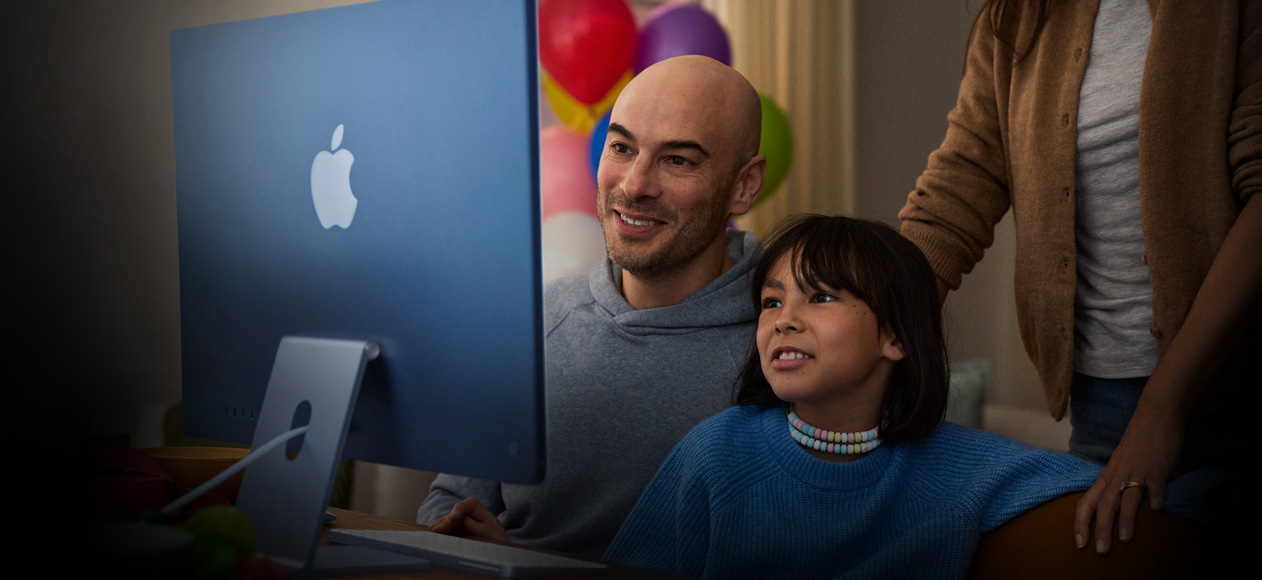 คนสองคนนั่งด้วยกันใช้ iMac โดยมีบุคคลที่สามยืนดูอยู่ด้านหลัง