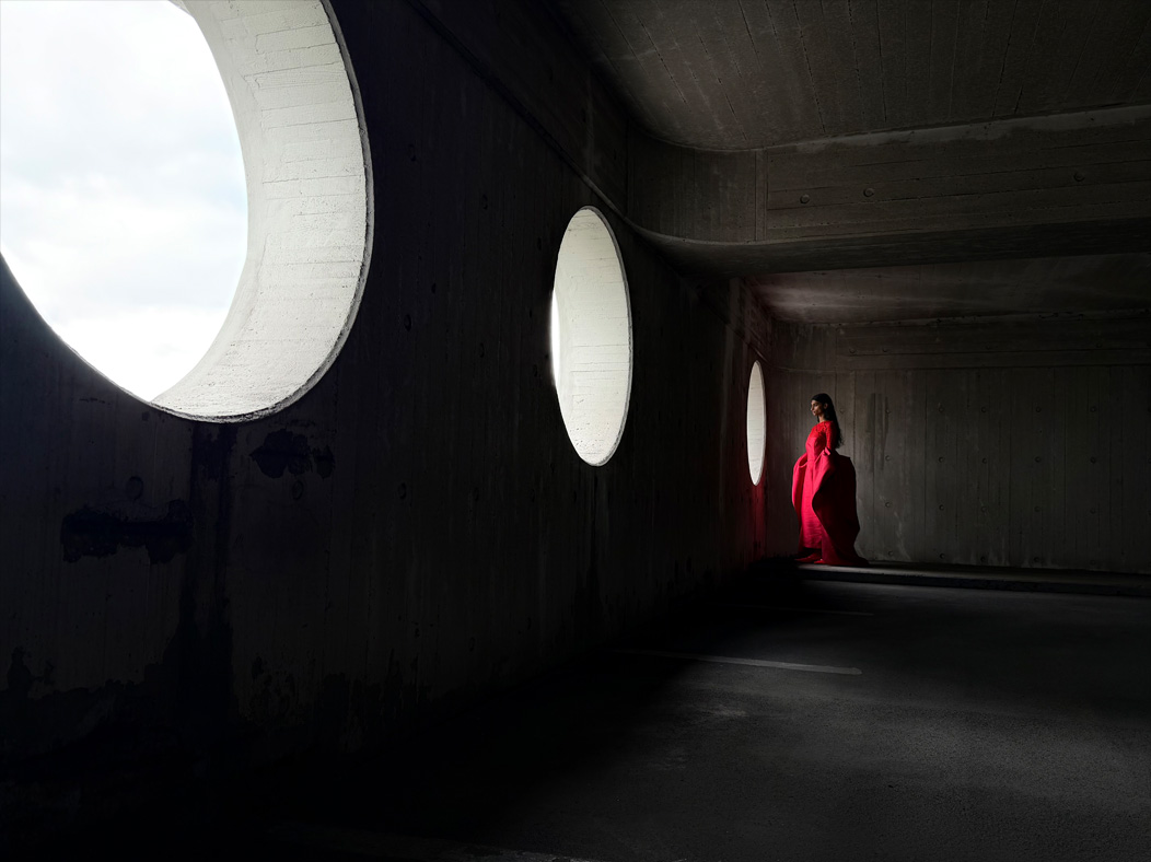 Een vrouw in een lange rode jurk wordt in een lege ruimte gefotografeerd. De Camera-app wisselt van brandpuntsafstanden om de mogelijkheden van het pro-camerasysteem te laten zien. Hier gekozen brandpuntsafstand: 28 mm